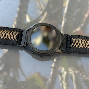May include: A black smartwatch with a black and gold paracord strap. The watch face is black and has a reflective surface. The strap has a buckle closure.