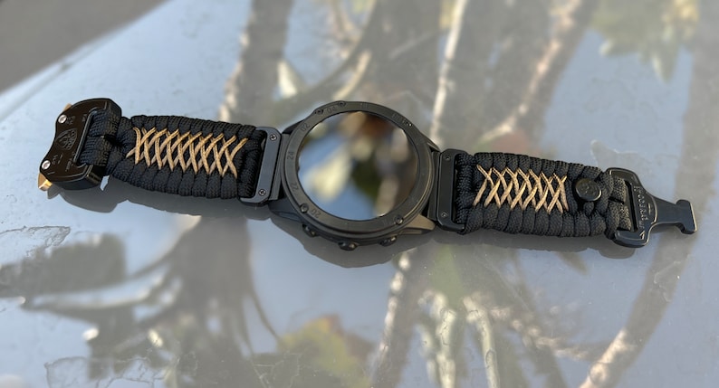 May include: A black smartwatch with a black and gold paracord strap. The watch face is black and has a reflective surface. The strap has a buckle closure.