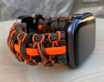 May include: A black Apple Watch with a camouflage and orange paracord band. The band is woven with a bright orange stripe.