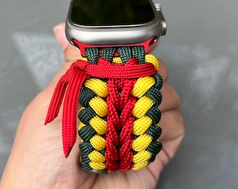 May include: A colorful watch band in green, yellow, and red, designed for a smartwatch. The band is made of woven nylon cord, with a braided pattern. The band is attached to a silver watch with a red accent.