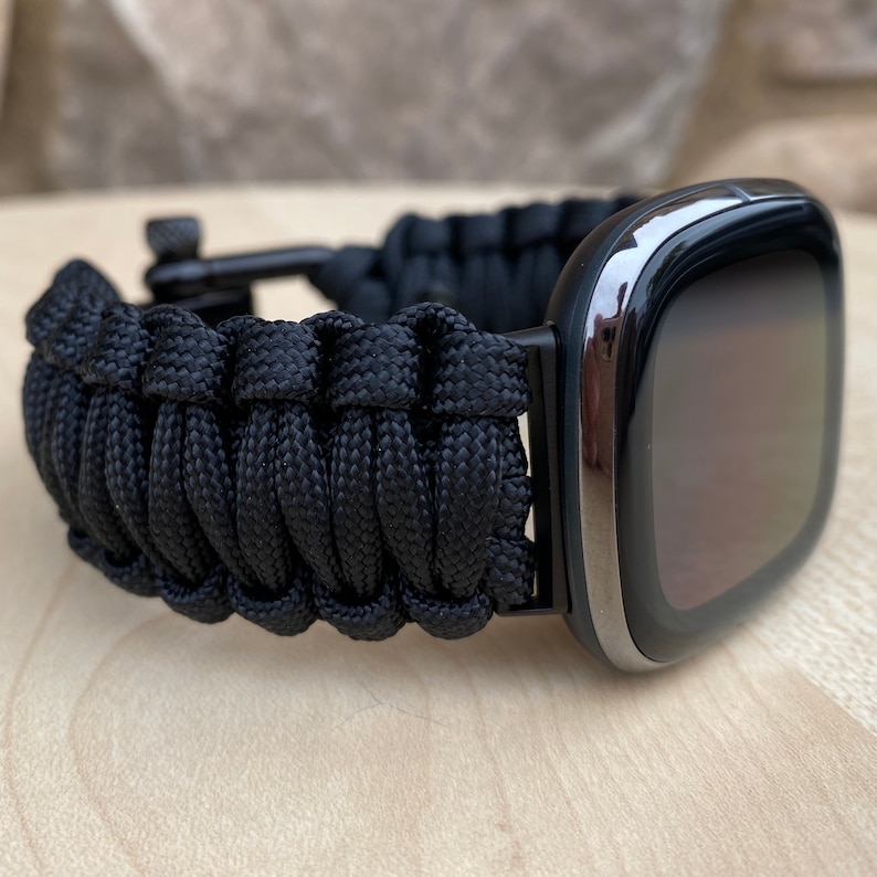 May include: Black paracord watch band with a black smartwatch attached. The band is made of woven paracord and has a metal buckle.