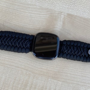 May include: A black smartwatch with a black paracord strap and metal clasps. The watch face is black and reflective.