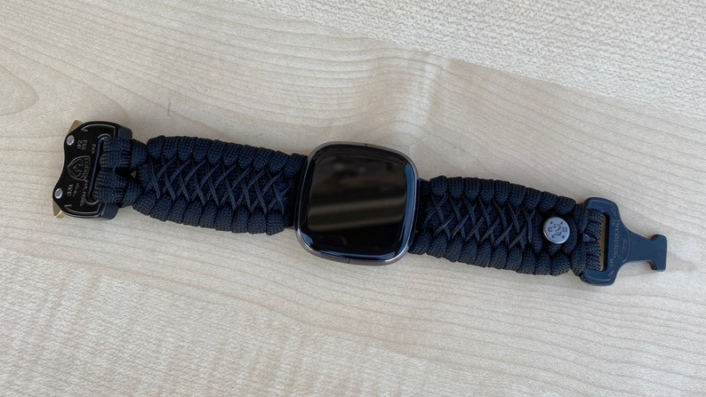 May include: A black smartwatch with a black paracord strap and metal clasps. The watch face is black and reflective.
