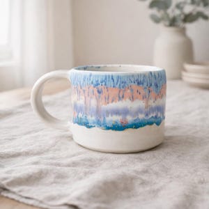 May include: White ceramic mug with a colourful, abstract design. The mug has a white base and handle, with a band of blue, pink, and purple hues around the middle. The mug sits on a neutral-coloured linen tablecloth.
