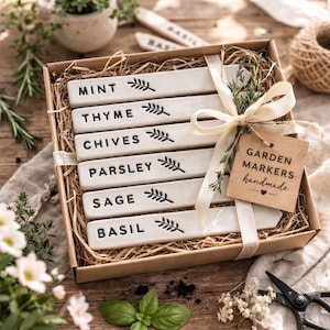 May include: A set of six rectangular white ceramic garden markers with black text reading Mint, Thyme, Chives, Parsley, Sage, and Basil, each featuring a small leaf icon. The markers are arranged in a brown cardboard box with straw filler.