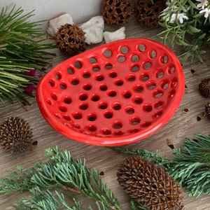 May include: A bright red ceramic soap dish with an irregular, rounded shape and a perforated surface featuring numerous circular holes throughout the base, designed with a smooth, glossy finish and a raised, slightly uneven outer rim.
