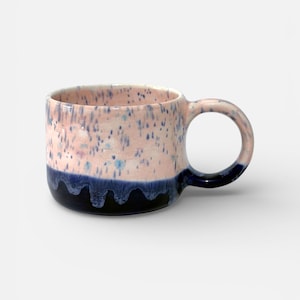 Può includere: Una tazza in ceramica con manico arrotondato. La tazza presenta una metà superiore rosa macchiettata e una metà inferiore blu scuro. Il manico e il bordo sono anch'essi blu scuro. La tazza è progettata per bevande calde.