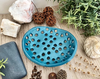 May include: A turquoise ceramic soap dish with numerous circular drainage holes. The dish is oval-shaped and sits on a light wood surface, surrounded by decorative elements like rocks, dried plants, and a bar of soap.