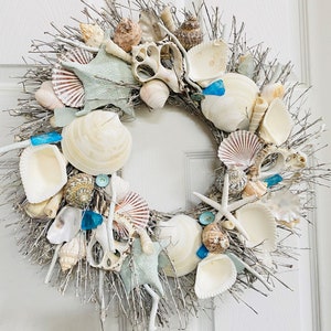 May include: A white twig wreath decorated with seashells, starfish, and blue glass beads. The wreath is a coastal-themed home decor item.