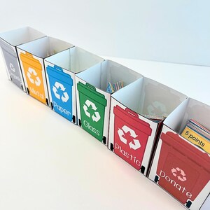May include: A set of six colorful cardboard bins with recycling symbols and labels for metal, paper, glass, plastic, donate, and 5 points. The bins are arranged in a row and are empty.