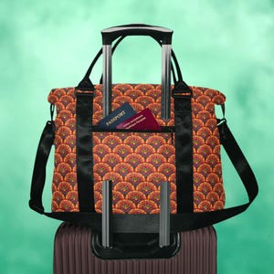 May include: A travel tote bag featuring a repeating orange, yellow, and dark blue fan-shaped pattern. The bag has black straps and a front pocket containing a blue passport and a red passport, mounted on the handle of a dark brown suitcase.