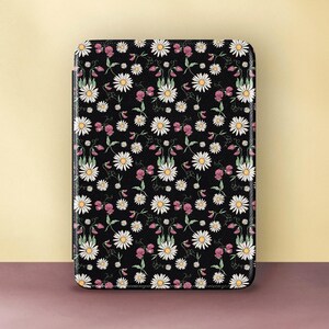 Könnte beinhalten: Eine rechteckige E-Reader-Hülle mit schwarzem Hintergrund und einem sich wiederholenden Muster aus weißen Gänseblümchen mit gelben Mitten, durchsetzt mit kleinen rosa Blütenknospen und grünen Stängeln mit dünnen, sich kräuselnden Ranken.