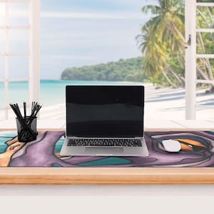 May include: A rectangular desk mat featuring a stylized purple and teal graphic print with a large orange eye motif. A silver laptop sits centered on the mat, accompanied by a white computer mouse and a black wire mesh pencil holder with pens.