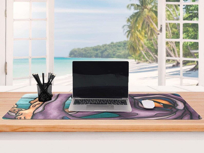 May include: A rectangular desk mat featuring a stylized purple and teal graphic print with a large orange eye motif. A silver laptop sits centered on the mat, accompanied by a white computer mouse and a black wire mesh pencil holder with pens.