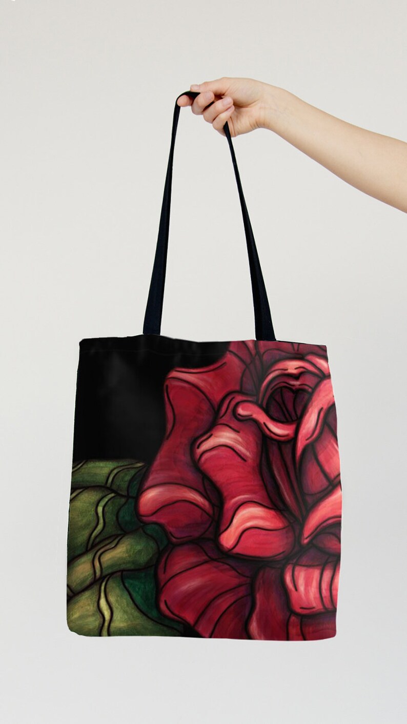 Op de afbeelding: Zwarte tote bag met een rode roos illustratie. De roos is getekend in een gestileerde, bijna cartoonachtige stijl. De roos is het hoofdelement van het ontwerp en neemt het grootste deel van de voorkant van de tas in.