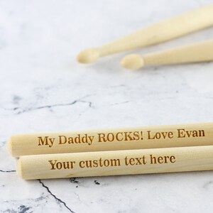 Puede incluir: Dos baquetas de madera con el texto "My Daddy ROCKS! Love Evan" y "Your custom text here" grabado en ellas.
