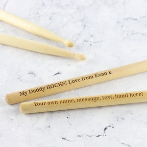 Puede incluir: Dos baquetas de madera con el texto "My Daddy ROCKS! Love from Evan x" y "Your own name, message, text, band here!" grabado en ellas.