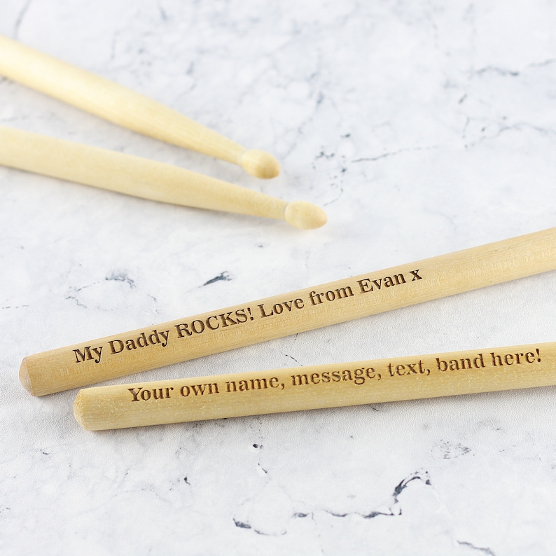Puede incluir: Dos baquetas de madera con el texto "My Daddy ROCKS! Love from Evan x" y "Your own name, message, text, band here!" grabado en ellas.