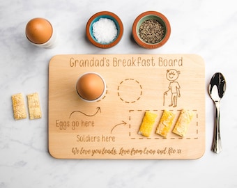 May include: A wooden breakfast board with the text "Grandad's Breakfast Board" engraved on it. The board has a place for an egg, a place for soldiers, and a drawing of a man with a cane. The text "Eggs go here", "Soldiers here", and "We love you loads, Love from Evan and Rio xx" is also engraved on the board.