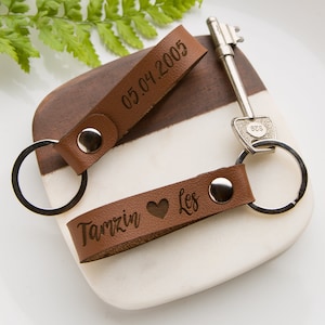May include: Two brown leather keychains with silver metal rings and snap buttons. One keychain is engraved with the date 05.04.2005, and the other is engraved with the text Tamzin heart Les. A silver metal key is attached to one of the keychains.