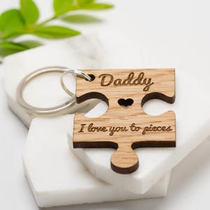 Op de afbeelding: Houten puzzelstukje sleutelhanger met het woord "Daddy" gegraveerd bovenaan en "I love you to pieces" onderaan. Een klein hartje is in het midden uitgesneden. De sleutelhanger is bevestigd aan een zilveren ring.