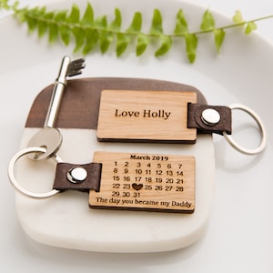 Może przedstawiać: Drewniany brelok do kluczy z dwoma grawerowanymi etykietami. Pierwsza etykieta głosi "Love Holly", a druga etykieta zawiera kalendarz na marzec 2019 z sercem oznaczającym 23 dzień i tekst "Dzień, w którym zostałeś moim tatą".