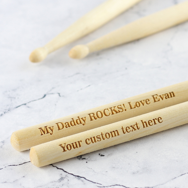 Puede incluir: Dos baquetas de madera con el texto "My Daddy ROCKS! Love Evan" y "Your custom text here" grabado en ellas.