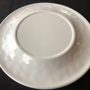 Op de afbeelding: Een witte keramische plaat met een licht golvende rand. De onderkant van de plaat heeft een verzonken midden met de tekst "Sur la table" en andere tekst. De plaat is rond en lijkt nieuw.