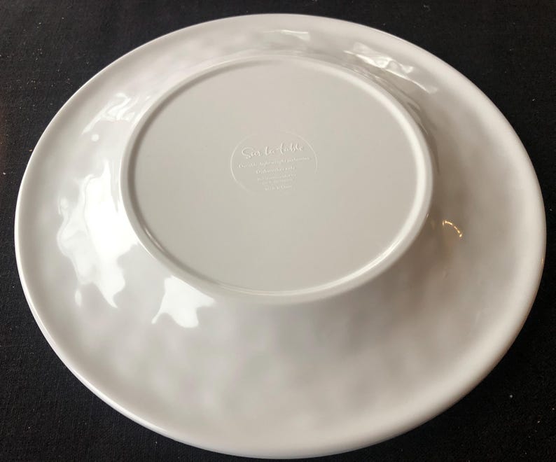 Op de afbeelding: Een witte keramische plaat met een licht golvende rand. De onderkant van de plaat heeft een verzonken midden met de tekst "Sur la table" en andere tekst. De plaat is rond en lijkt nieuw.