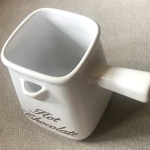 May include: White ceramic hot chocolate pot with a handle and the words "Hot Chocolate" written on the side.