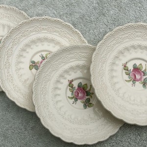 May include: Set of four white ceramic saucers with a delicate lace-like design and a pink rose floral pattern in the center.