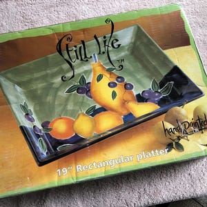 May include: A rectangular 19-inch platter in a box featuring a Still Life brand logo. The platter is decorated with a hand-painted design of yellow pears, oranges, and dark purple grapes against a green and black background. The box displays the text Still Life, hand painted, and 19 inch Rectangular platter.