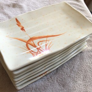 May include: A stack of rectangular ceramic plates with a cream-colored base, featuring a hand-painted orange grass motif on the surface. The plates have a subtle speckled pattern in light orange and slightly curved, raised edges for a defined shape.