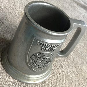 May include: A metallic gray tankard featuring a raised shield emblem on the front. The emblem displays the text VIRGINIA TECH above a circular seal that reads VIRGINIA POLYTECHNIC INSTITUTE AND STATE UNIVERSITY. The mug has a sturdy handle.