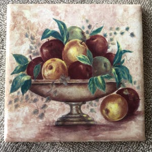 May include: Square ceramic tile featuring a still life of apples in a decorative bowl. The apples are various colors, including red, yellow, and green, with green leaves. The bowl is a light brown color, and the background is a soft pink.