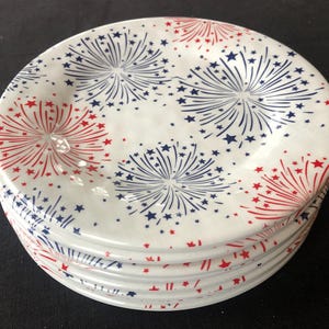 Op de afbeelding: Een stapel witte keramische borden met een vuurwerkontwerp. Elk bord heeft rode en blauwe uitbarstingen met steraccenten, waardoor een patriottisch thema ontstaat. De borden zijn op een donkere ondergrond gestapeld.