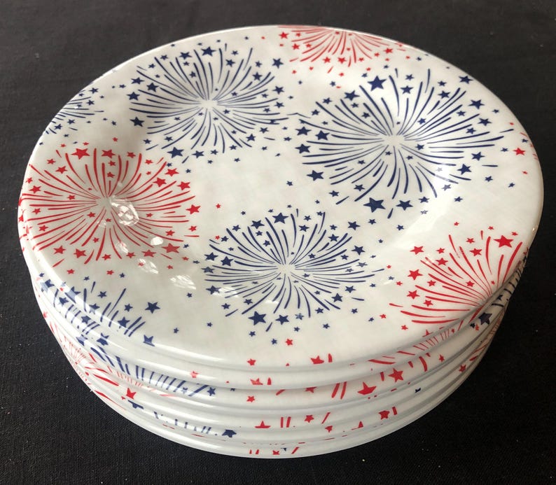 Op de afbeelding: Een stapel witte keramische borden met een vuurwerkontwerp. Elk bord heeft rode en blauwe uitbarstingen met steraccenten, waardoor een patriottisch thema ontstaat. De borden zijn op een donkere ondergrond gestapeld.