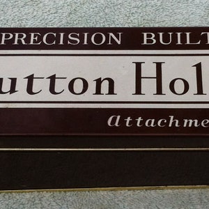 May include: A rectangular brown metal box with a white and brown label. The label reads "PRECISION BUILT Button Holer Attachment" in a serif font. The box has a gold trim.