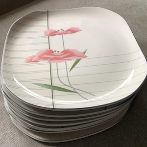 May include: A stack of ten white ceramic dinner plates with a floral design. The plates have a raised rim and a slightly squared shape. The floral design features pink flowers with green leaves on a white background with gray lines.