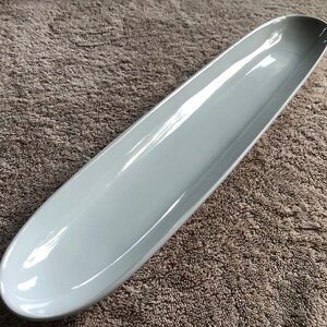May include: A long, narrow, oval-shaped serving dish with a smooth, glossy light gray finish. The dish features a shallow, elongated basin with rounded edges and a continuous, curved rim, presenting a clean and simple design for food presentation.