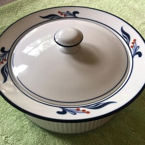 May include: White ceramic casserole dish with a lid. The dish has a navy blue rim and a decorative pattern of navy blue leaves and red berries. The lid has a white knob and a navy blue rim. The dish is round and has a ribbed exterior.