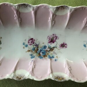 May include: A vintage oval-shaped serving dish with a pink and white floral design. The dish has a scalloped edge and is decorated with gold accents.