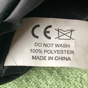 May include: A white tag with black text that reads "CE" and "Do Not Wash 100% Polyester Made in China". The tag is attached to a black fabric.