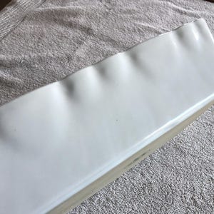 May include: A rectangular, white ceramic baking dish with a slightly wavy edge. The dish has a glossy finish and appears to be clean. The background is a light gray, textured surface. This dish is suitable for baking and serving.