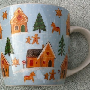 May include: A blue ceramic mug with a winter village scene. The mug features gingerbread houses, snowmen, trees, and horses. The mug has a white handle.