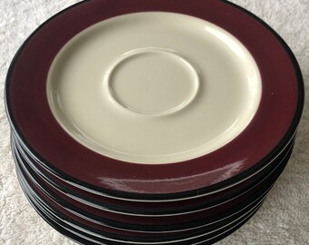 May include: A stack of six saucer-shaped plates with a burgundy rim and a black trim. The plates are made of ceramic and have a white center.