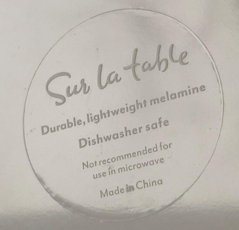 Op de afbeelding: Een helder, cirkelvormig etiket met de woorden "Sur la table" in een cursief lettertype. Het etiket bevat ook de tekst "Durable, lightweight melamine, Dishwasher safe, Not recommended for use in microwave, Made in China."