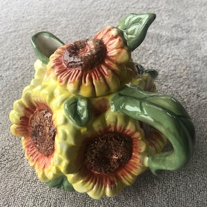 May include: A ceramic teapot shaped like a cluster of sunflowers. The teapot features yellow petals with red tips, brown centers, and green leaves and handle. The teapot is a decorative kitchen item.