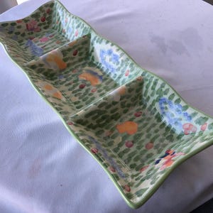 May include: A ceramic serving dish with three compartments and a wavy design. The dish is painted with a green background and a pattern of small green brushstrokes. It features colorful floral accents in orange, blue, and pink.