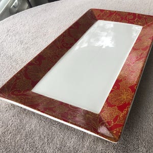 May include: Rectangular serving platter with a white center and a red and gold paisley patterned border. The dish has a glossy finish and is likely made of ceramic or porcelain. Ideal for serving appetizers or desserts.